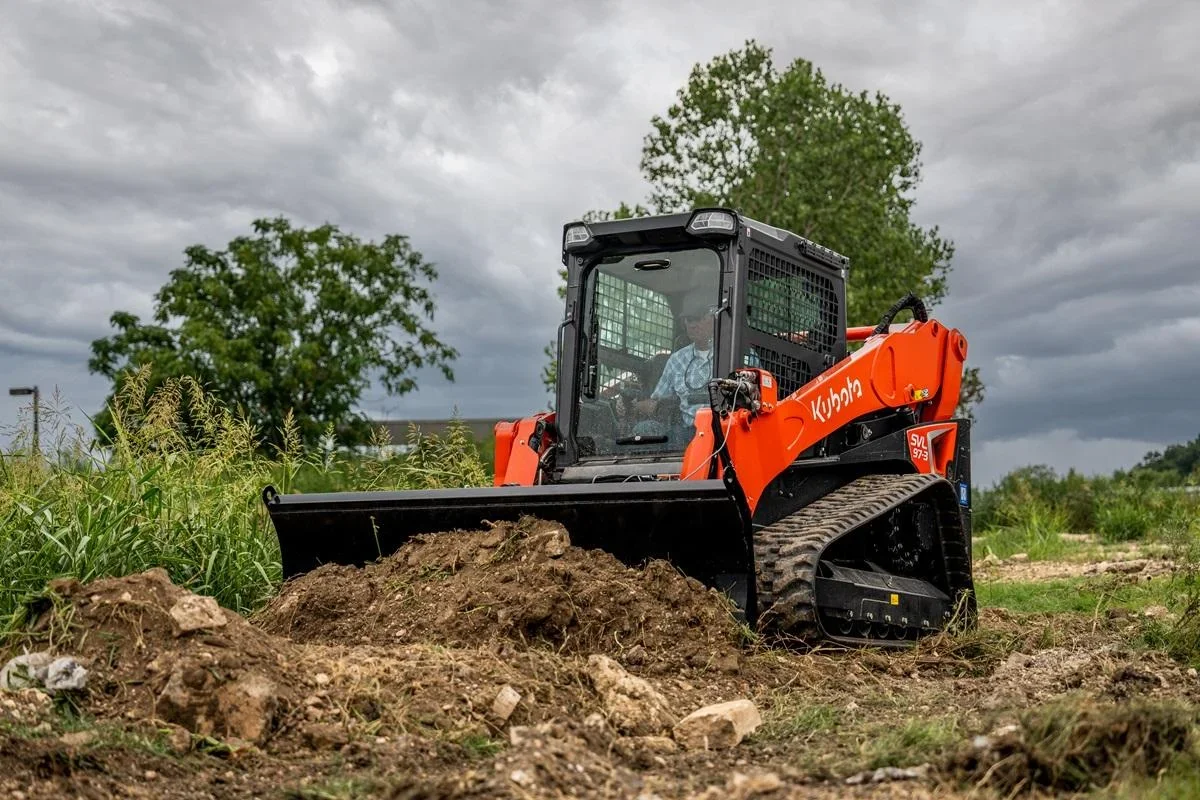 Orange Kubota compact track loader with black bucket grading loose soil and rocks on a cleared site